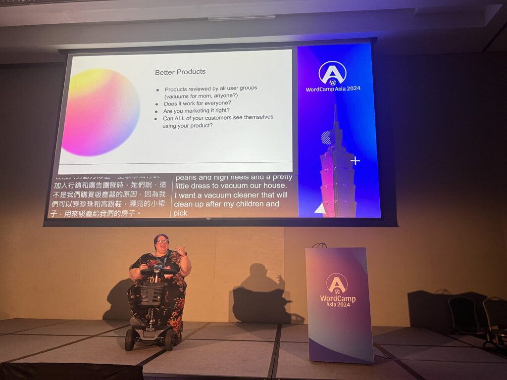 Michelle Frechette on the stage at WC Asia. She's siting on a mobility scooter. Above her is a screen showing her slides. The slide on display discusses how inclusion makes products better.