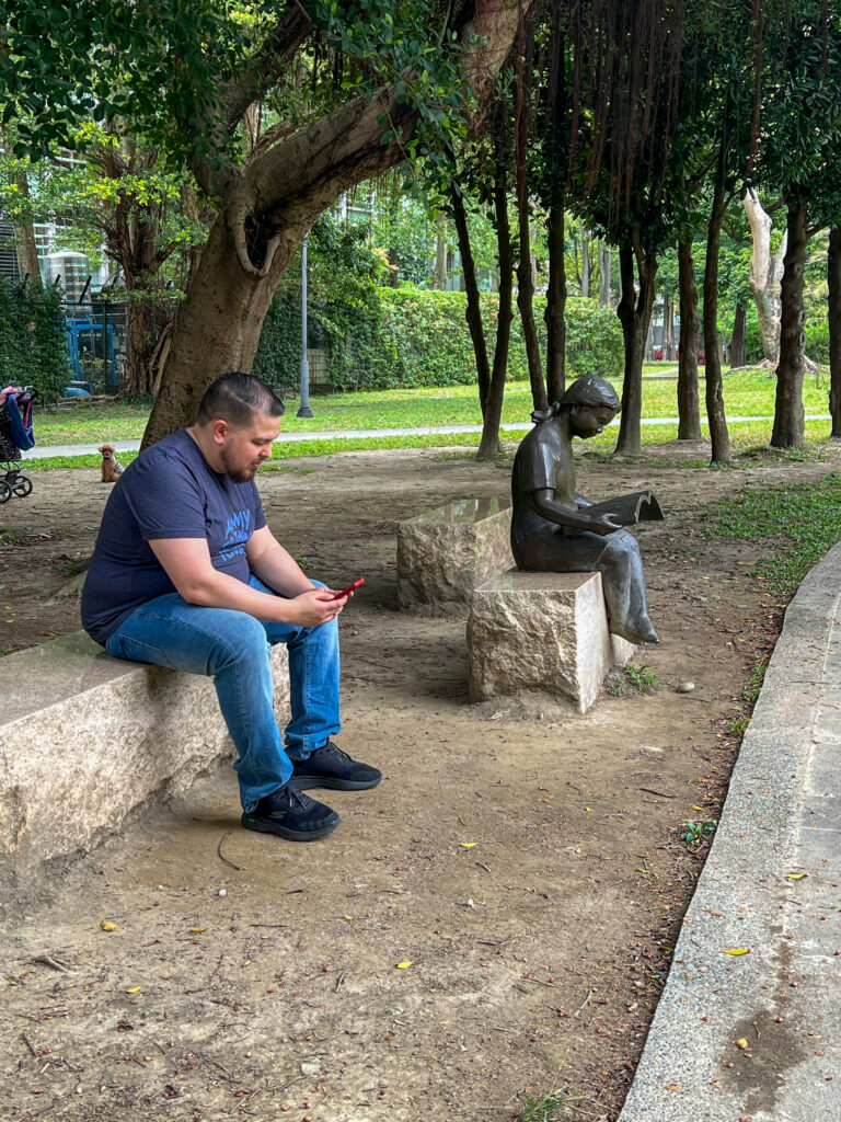 Jeff Betancourt replicates art by reading on his phone on a bench near a sculpture of someone reading a book on a bench