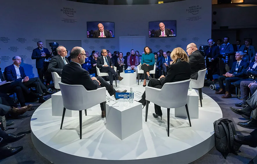 A group of people seated in a circular arrangement on a stage at the World Economic Forum, with an audience observing. The setting includes screens displaying speakers and a central table with nameplates and water bottles.