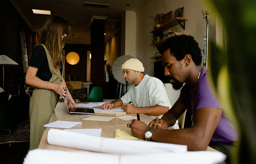 Three people working at a desk in an office setting, with one person standing and two sitting, engaged in writing and discussion.