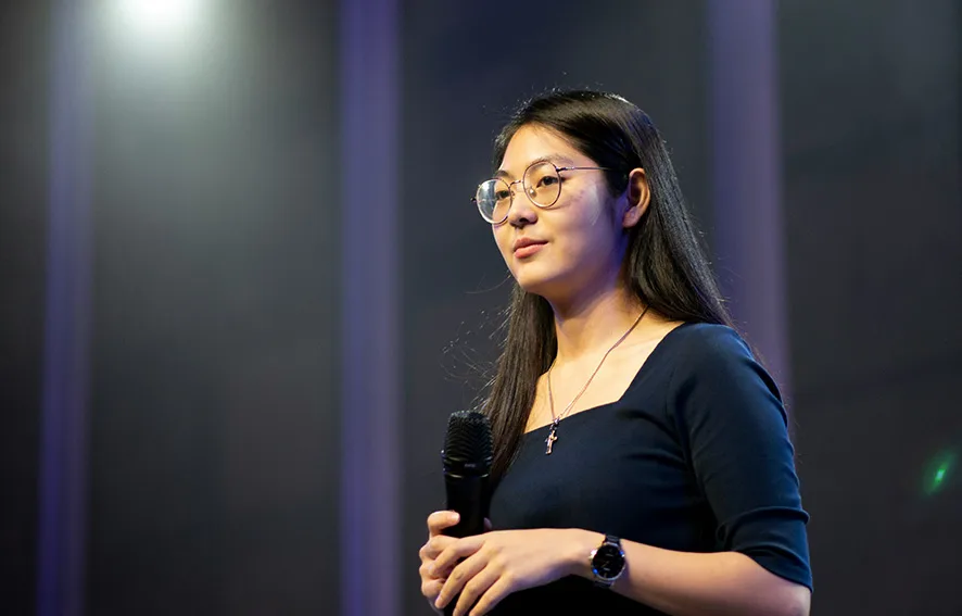 A woman with long hair and eyeglasses is holding a microphone while standing on a stage, wearing a dark top.