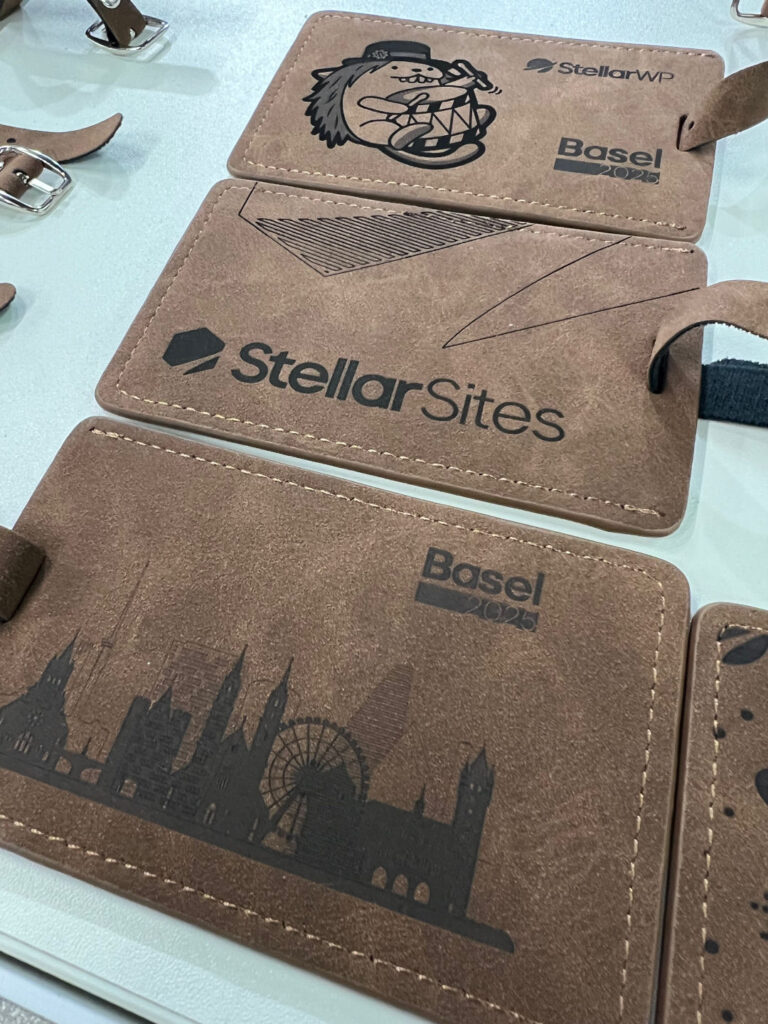 Brown leather luggage tags featuring printed designs, including a cartoon figure with a drum, the text 'StellarWP' and 'Basel 2025,' the logo 'StellarSites,' and silhouettes of buildings with a Ferris wheel.