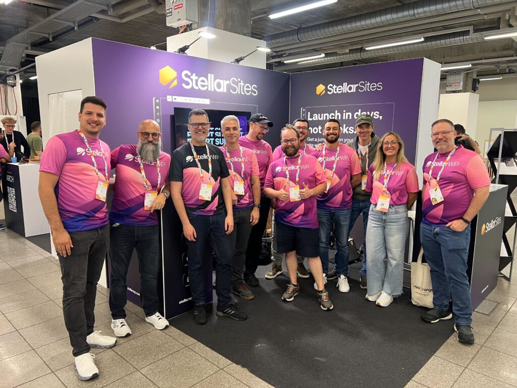 A group of people wearing matching pink and purple "StellarWP" shirts stand together in front of a booth labeled "StellarSites" at an event. They are smiling and appear to be part of a team presenting or representing their company.