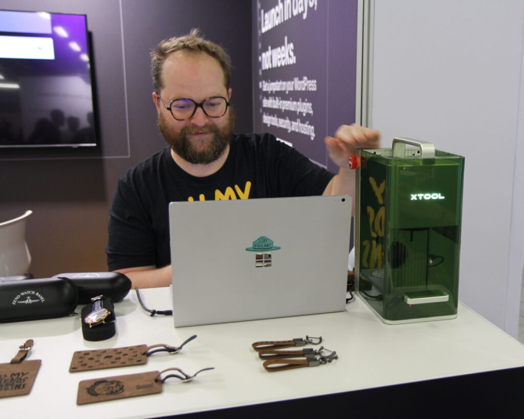 A man with glasses and a beard working at a table with a laptop and an xTool laser engraver. Various leather crafts and accessories are displayed on the table.