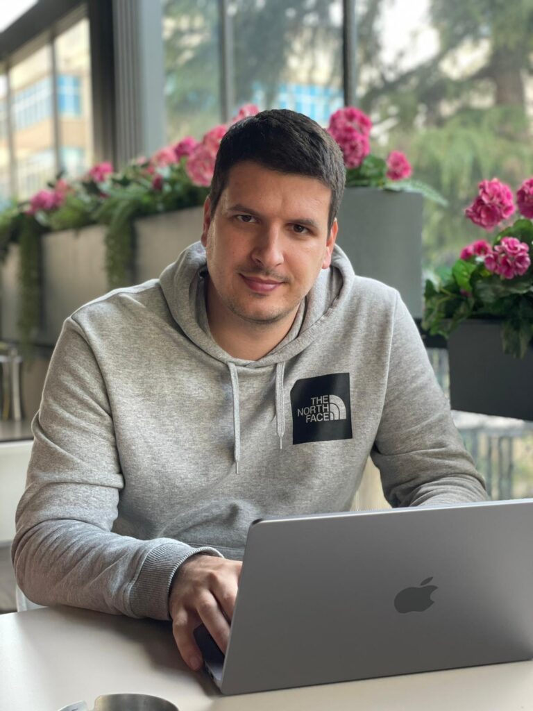 A person in a gray hoodie working on a laptop with pink flowers in the background.