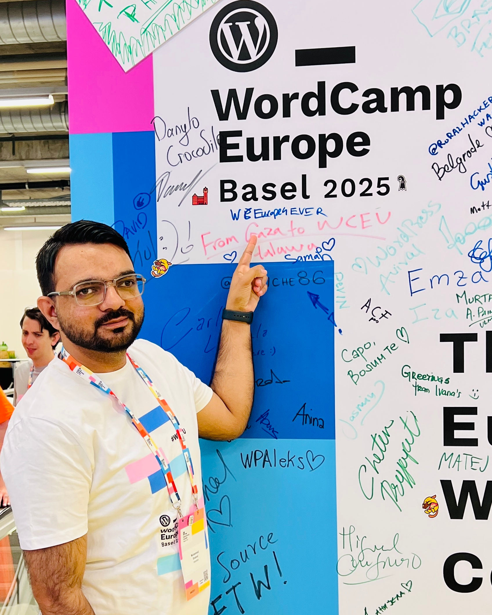 A person wearing a WordCamp Europe Basel 2025 t-shirt stands next to a colorful wall with various handwritten messages and signatures. They are pointing at a specific message on the wall. The background shows a blurred person and industrial ceiling details.