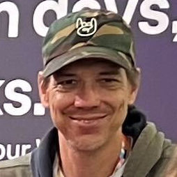 Person wearing a camouflage cap with a white graphic of a hand making a rock sign, smiling in front of a blurred background with text.