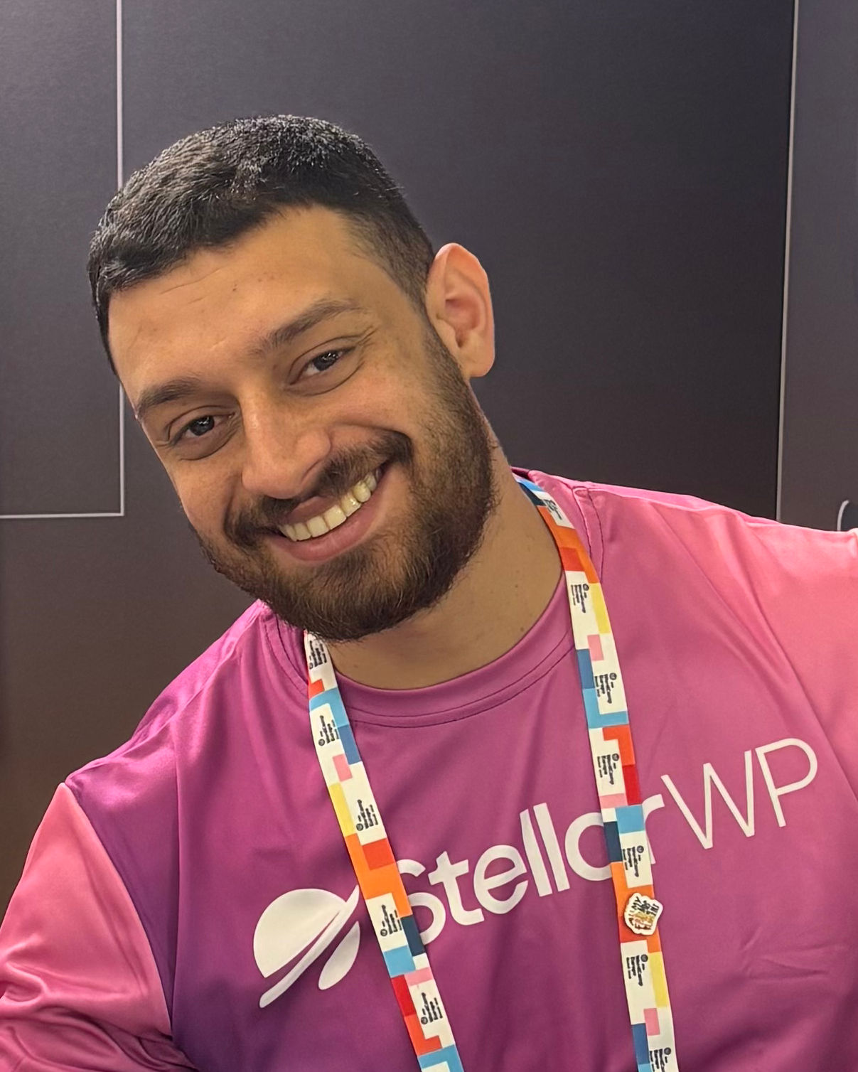 A man with short hair and a beard is smiling at the camera. He is wearing a bright pink shirt with the logo and text 'StellarWP' visible, and a colorful lanyard around his neck. The background is dark and blurred.