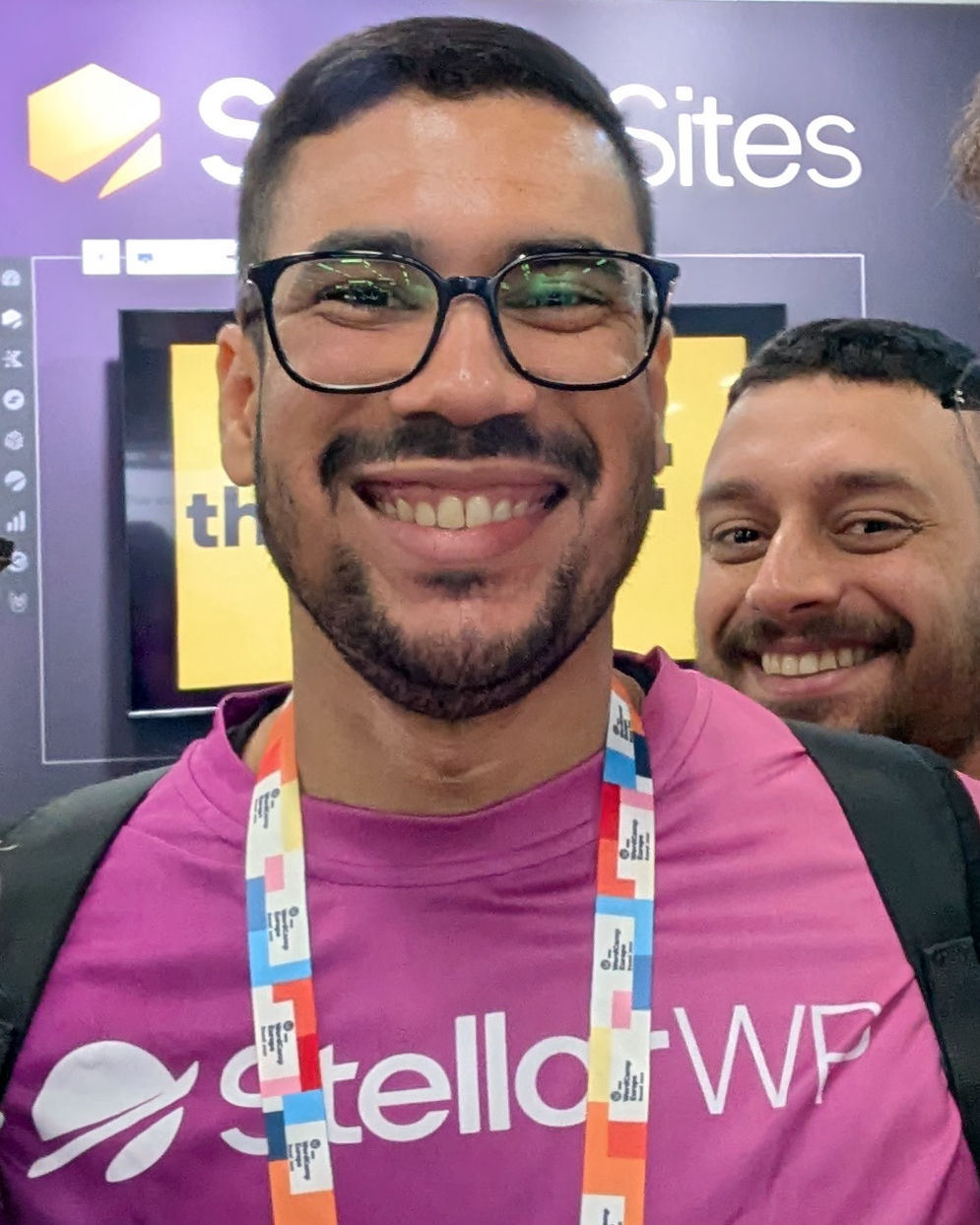 Two men smiling; the one in front is wearing glasses and a pink "stellarWP" shirt with a colorful lanyard, standing in front of a purple backdrop with partial text visible.