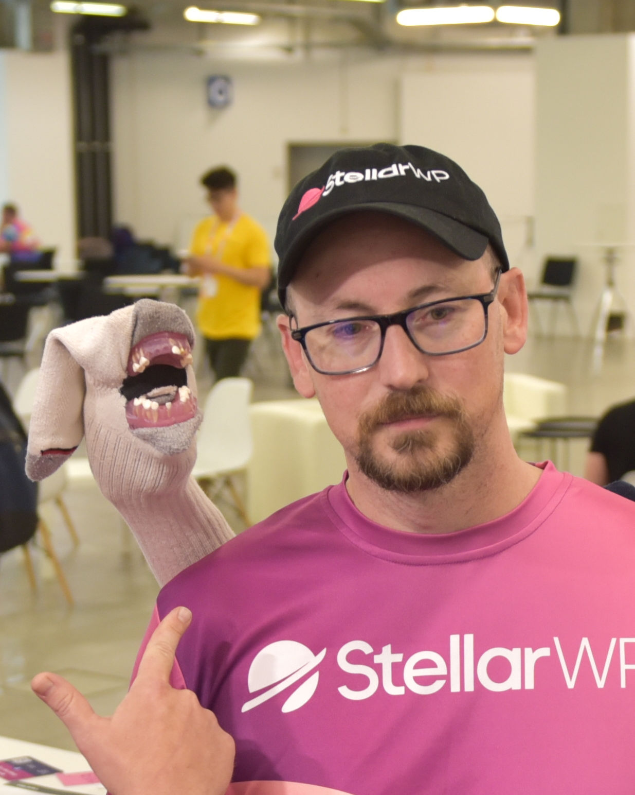 A man wearing glasses, a black cap with 'StellarWP,' and a pink shirt with the same logo, is holding a sock puppet with an open mouth showing teeth. The setting is an indoor event space with blurred people and chairs in the background.