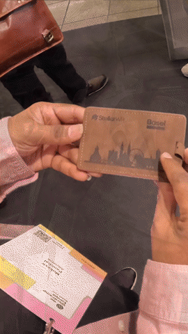 A person in a pink sweater holds a leather card holder with a city skyline embossed on it and the words "Stark WiFi" and "Basel."