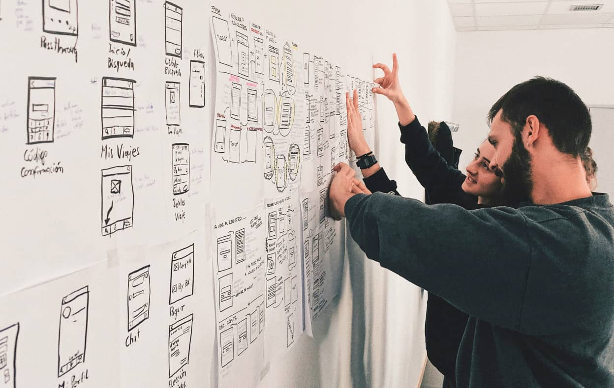 Two people discussing and pointing at sketches and wireframes on a wall, which feature drawn designs and Spanish text related to mobile app interfaces.