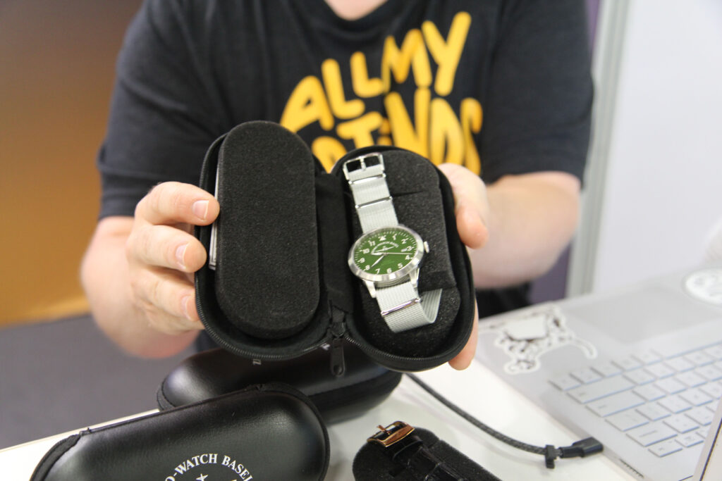 A person holding an open black watch case with a green-faced watch inside, featuring silver hands and a grey strap. The person is wearing a t-shirt with bright yellow text, and there is a partially visible laptop on the table nearby.