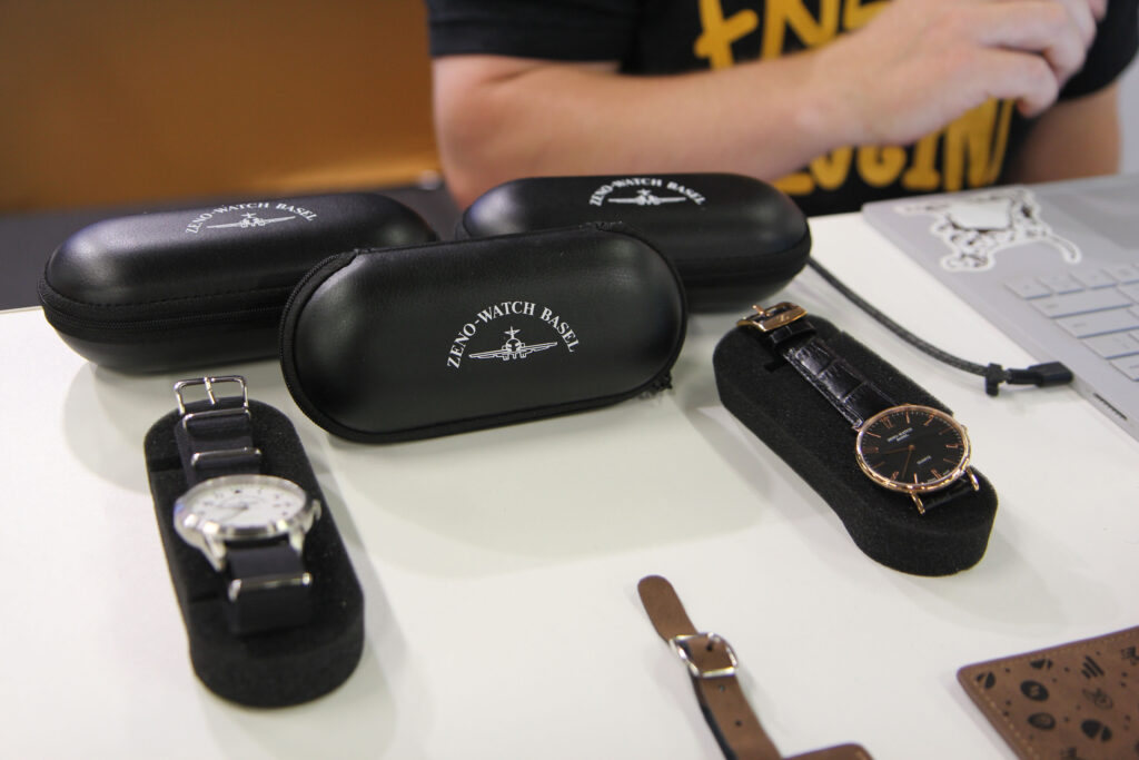Two watches displayed in foam holders on a table, accompanied by several black leather cases labeled 'Zeno-Watch Basel'; a person's arm and a laptop are visible in the background.