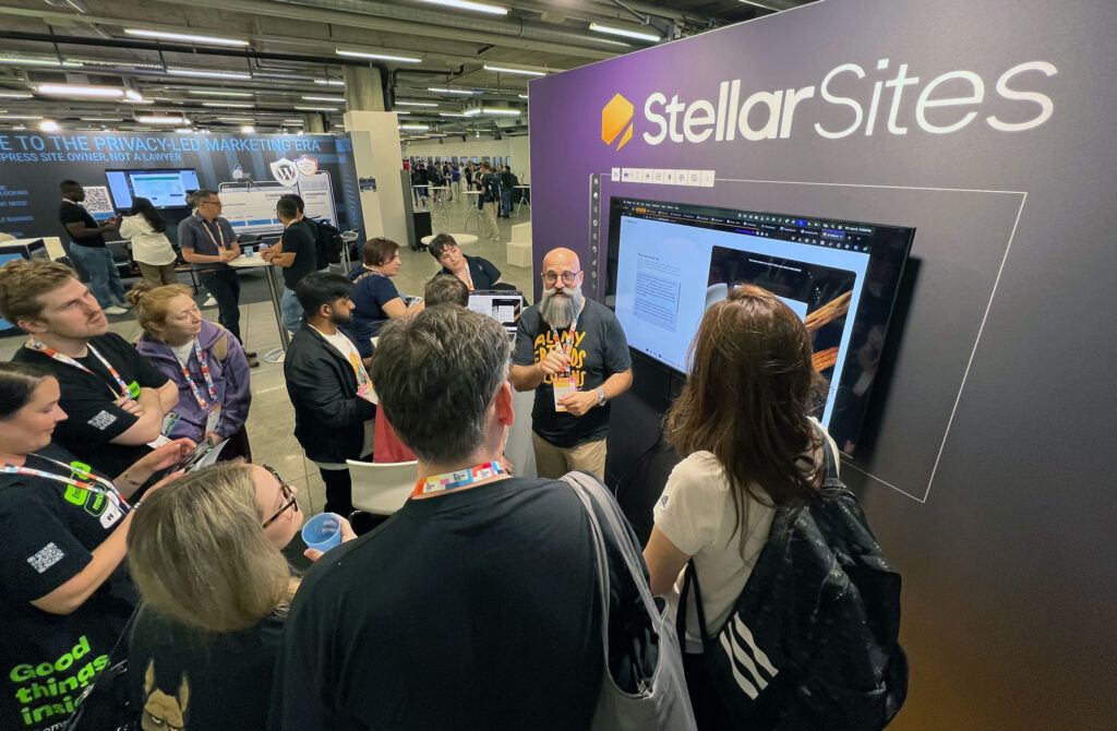 A presenter at the StellarSites booth demonstrates a product to a group of attendees at a tech conference. The backdrop has the StellarSites logo, and the setting is a large hall with other exhibitors in the background. Attendees appear engaged, some with lanyards and taking notes.