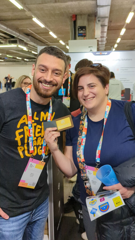 Two people smiling at a conference, with one person holding a "Winner" card and both wearing colorful lanyards with badges.