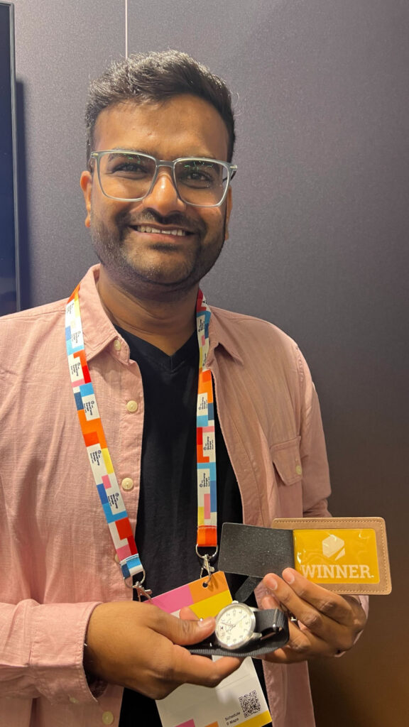 A man wearing glasses and a pink shirt, holding a watch and a card labeled 'Winner', with a colorful lanyard around his neck.